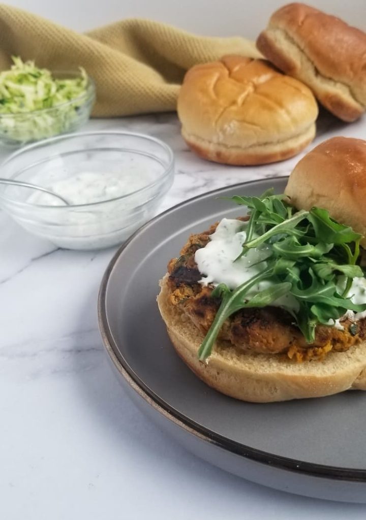 plate with one zucchini turkey burger on a bun topped with arugula and tzatziki sauce, small bowl of shredded zucchini, white dipping sauce and extra buns in the background