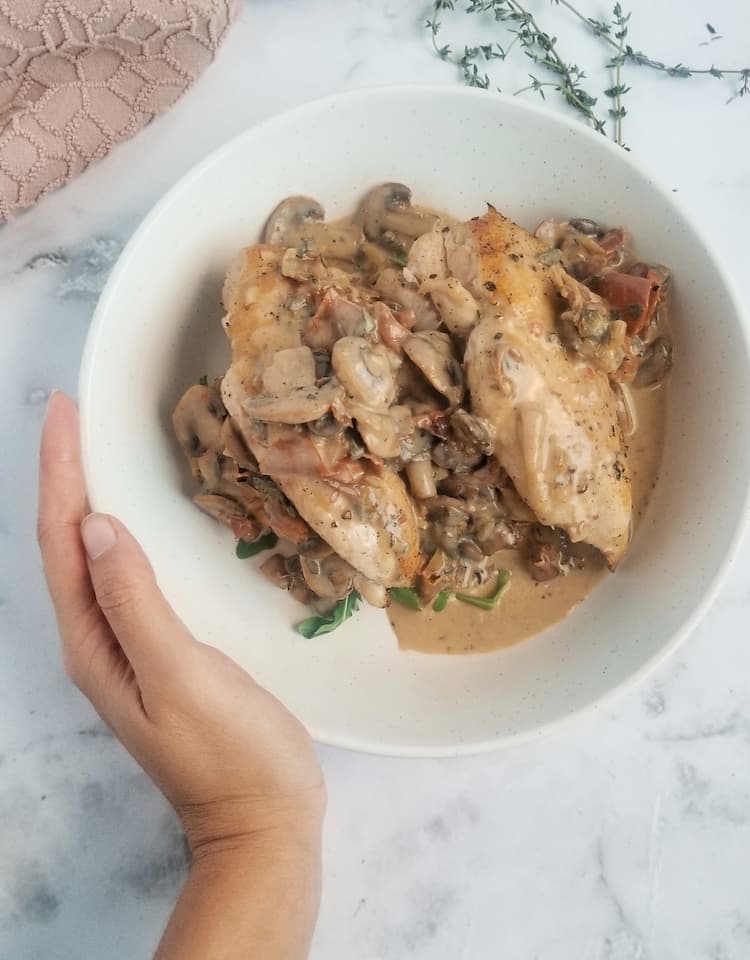 hand touching a bowl of two stuffed chicken breasts with creamy mushroom sauce around and on top