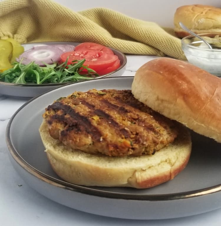 plain zucchini turkey burger on a bottom bun, top bun leaning against it, with a plate of toppings (pickles, tomatoes, red onions, arugula) in the background with an extra bun and a small bowl of a white dipping sauce with a spoon