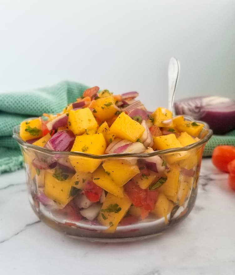 jar of mango habanero salsa - side profile of a small glass bowl full of chopped mangoes, red onions, parsley, red peppers, habaneros in the back ground and half a red onion, spoon sticking out of the bowl