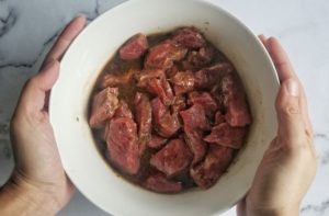 hands holding a bowl of steak strips marinating in a brown sauce