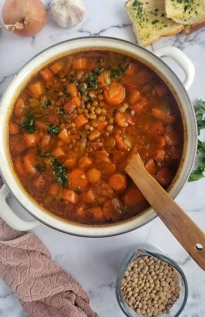 big pot of lentil soup with parsley, carrots, tomatoes, a whole onion and bulb of garlic in the background, a cup of uncooked lentils and a bunch of fresh parsley on the side, wooden spoon in the pot, 2 slices of garlic bread on the side