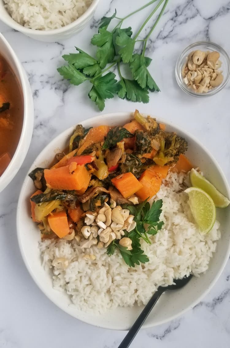 bowl of cubed sweet potato with other veggies such as spinach and sliced mushrooms, rice, parsley, chopped cashews and sliced lime, spoon in the bowl, fresh bunch of parsley and bowl of rice in the background, small bowl of cashews in the background