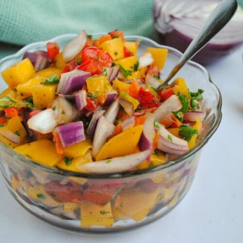 chunks of mango, red onion, parsley, red pepper and habaneros in a small glass bowl, side to top profile, 2 habaneros on the side and a half a red onion