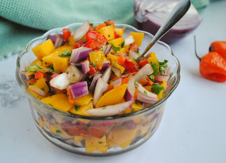 chunks of mango, red onion, parsley, red pepper and habaneros in a small glass bowl, side to top profile, 2 habaneros on the side and a half a red onion