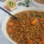 big bowl of lentil soup with carrots, tomatoes, onions and parsley, 2 slices of garlic bread in the background, with another bowl of lentil soup and a whole onion and some parsley