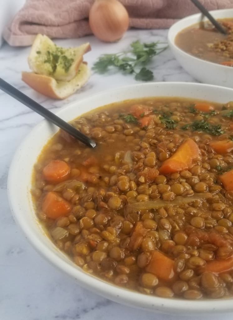 big bowl of lentil soup with carrots, tomatoes, onions and parsley, 2 slices of garlic bread in the background, with another bowl of lentil soup and a whole onion and some parsley