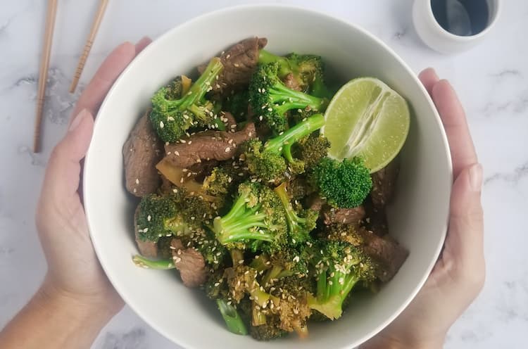 hands holding a bowl of beef and broccoli, half a lime, soy sauce and chopsticks in the background