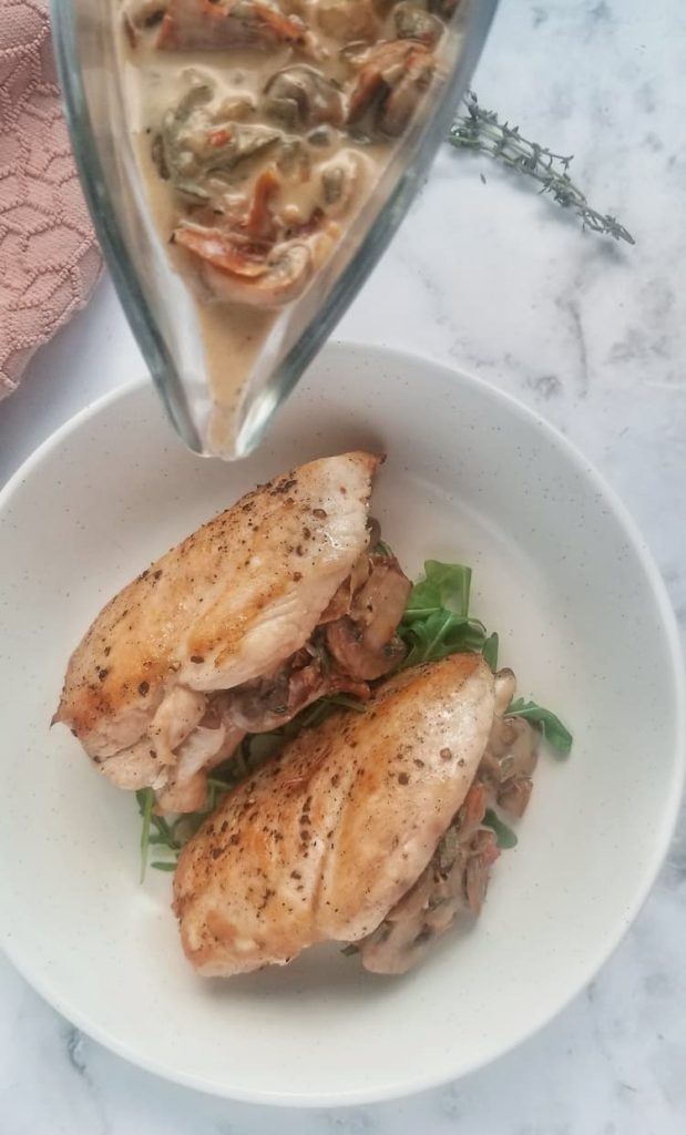 a gravy boat with prosciutto mushroom cream sauce pouring over two seasoned chicken breasts stuffed with the same sauce