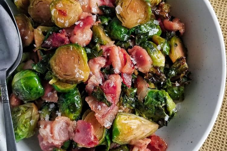 a big bowl of cooked brussel sprouts baked with bacon and maple syrup finished with maldon sea salt flakes, two spoons in the bowl, bowl on a yellow cloth