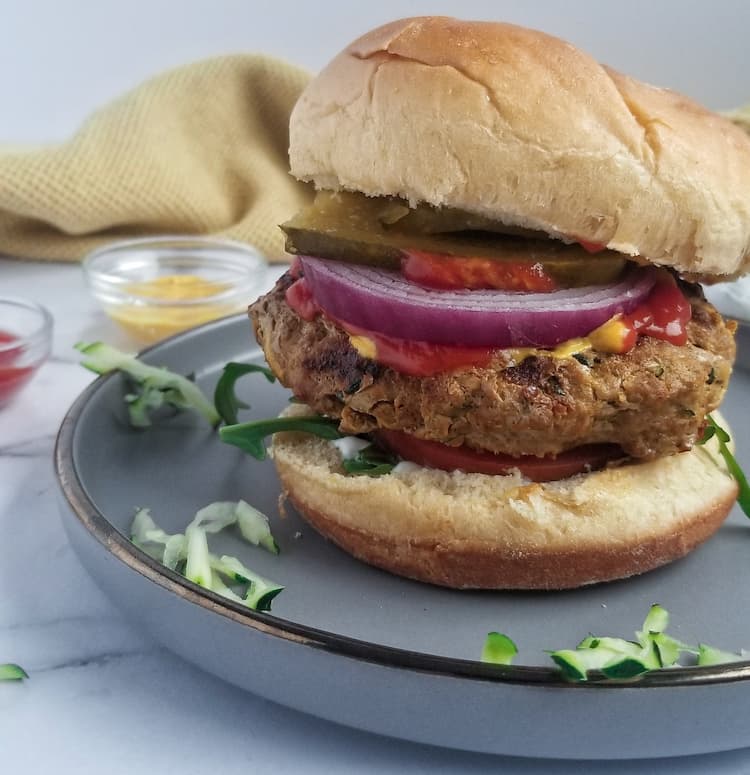 a zucchini turkey burger on a bun topped with pickles, mustard, ketchup, arugula, tomato, red onion, mustard and ketchup in the background, shredded zucchini pieces around