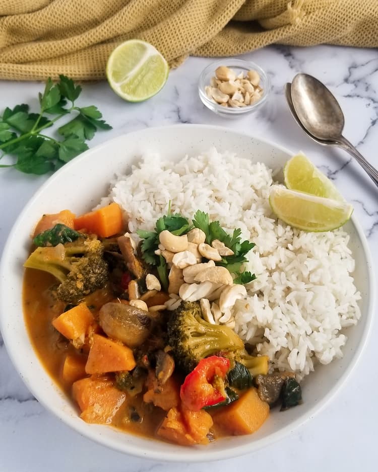 bowl of sweet potato curry with chopped parsley, rice and cashews, half a lime, bunch of parsley and cashews in the background