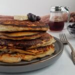 stack of ricotta pancakes with blueberries with 3 fresh blueberries and a rectangle piece of butter, bowl of blueberries in the back, syrup in a container in the background, fork next to plate