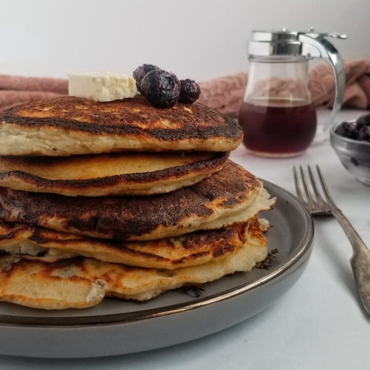 stack of ricotta pancakes with blueberries with 3 fresh blueberries and a rectangle piece of butter, bowl of blueberries in the back, syrup in a container in the background, fork next to plate