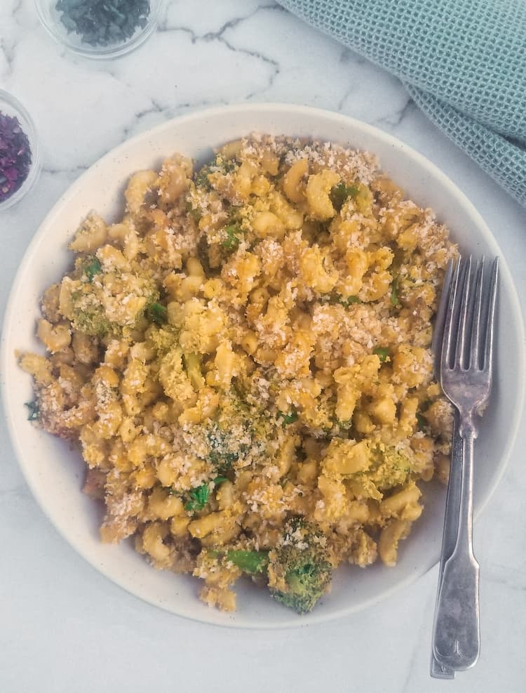 larger white bowl full of vegan mac and cheese baked with broccoli and breadcrumbs, two forks in the bowl, two small dishes of fresh chopped parsley and chilli flakes in the background