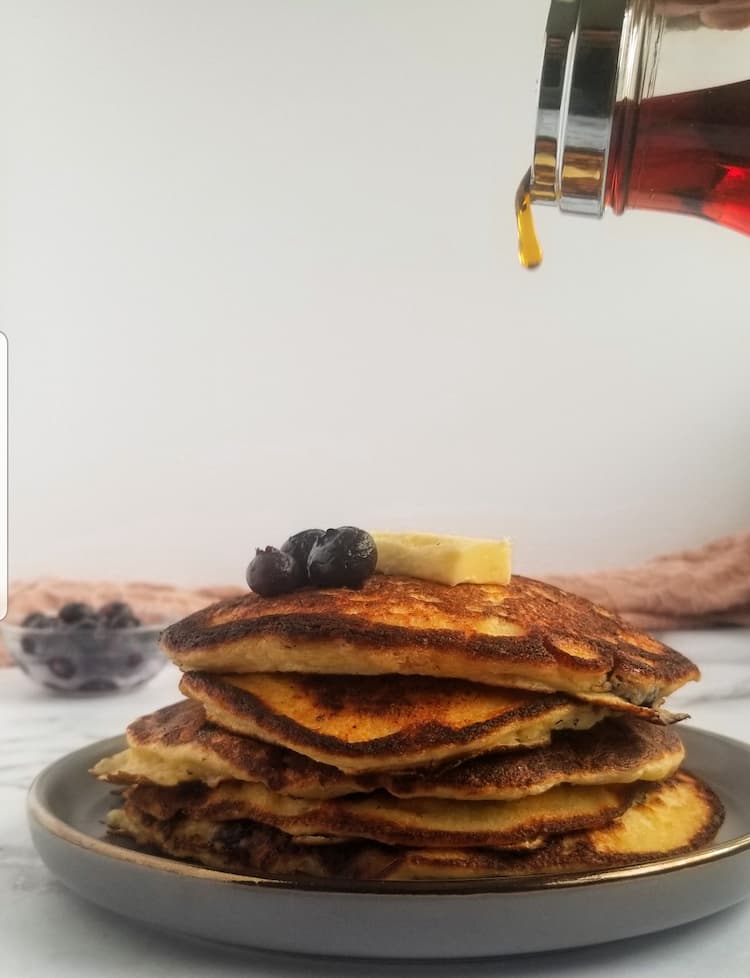 stack of ricotta pancakes with blueberries, fresh bowl of blueberries in the background and on top, block of butter on top, maple syrup dripping out of a container on the top of frame