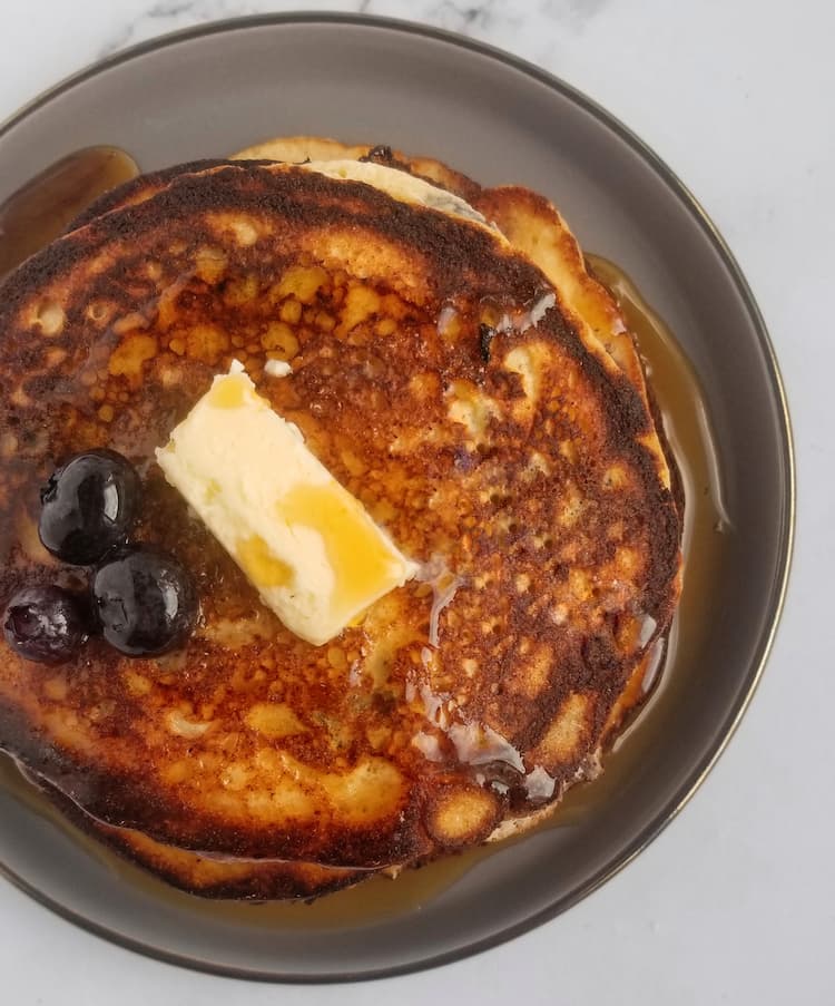 stack of ricotta pancakes with blueberries with 3 fresh blueberries and a rectangle piece of butter, sitting in maple syrup