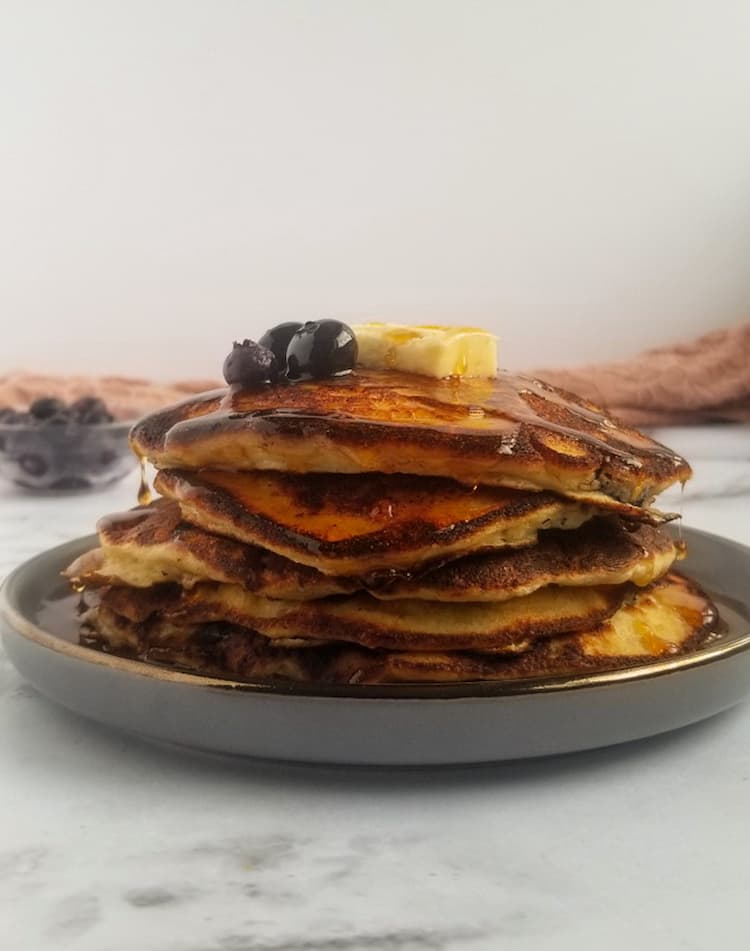 stack of ricotta pancakes with blueberries with 3 fresh blueberries and a rectangle piece of butter, bowl of blueberries in the back, syrup dripping off the sides and top of the pancakes