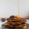 stack of ricotta pancakes with blueberries with 3 fresh blueberries and a rectangle piece of butter, bowl of blueberries in the back, syrup being poured onto the pancakes