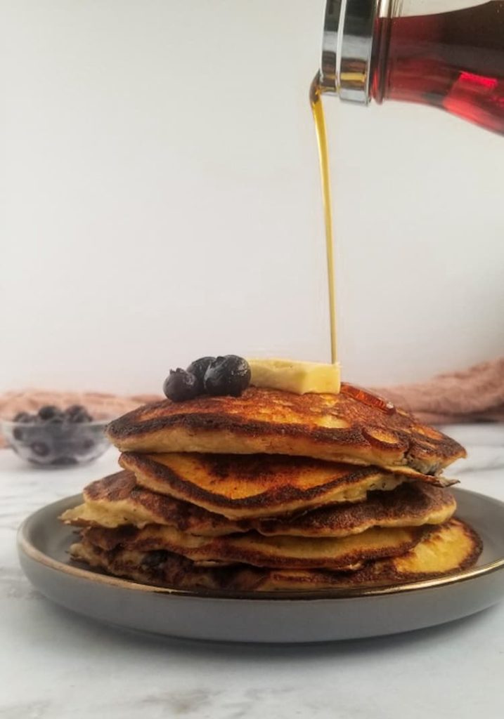 stack of ricotta pancakes with blueberries with 3 fresh blueberries and a rectangle piece of butter, bowl of blueberries in the back, syrup being poured onto the pancakes