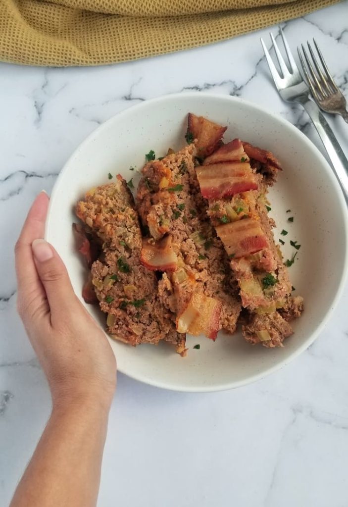 hand holding bowl with 3 slices of keto meatloaf topped with cooked bacon and fresh chopped parsley, two silver forks on the right side, yellow cloth in the background