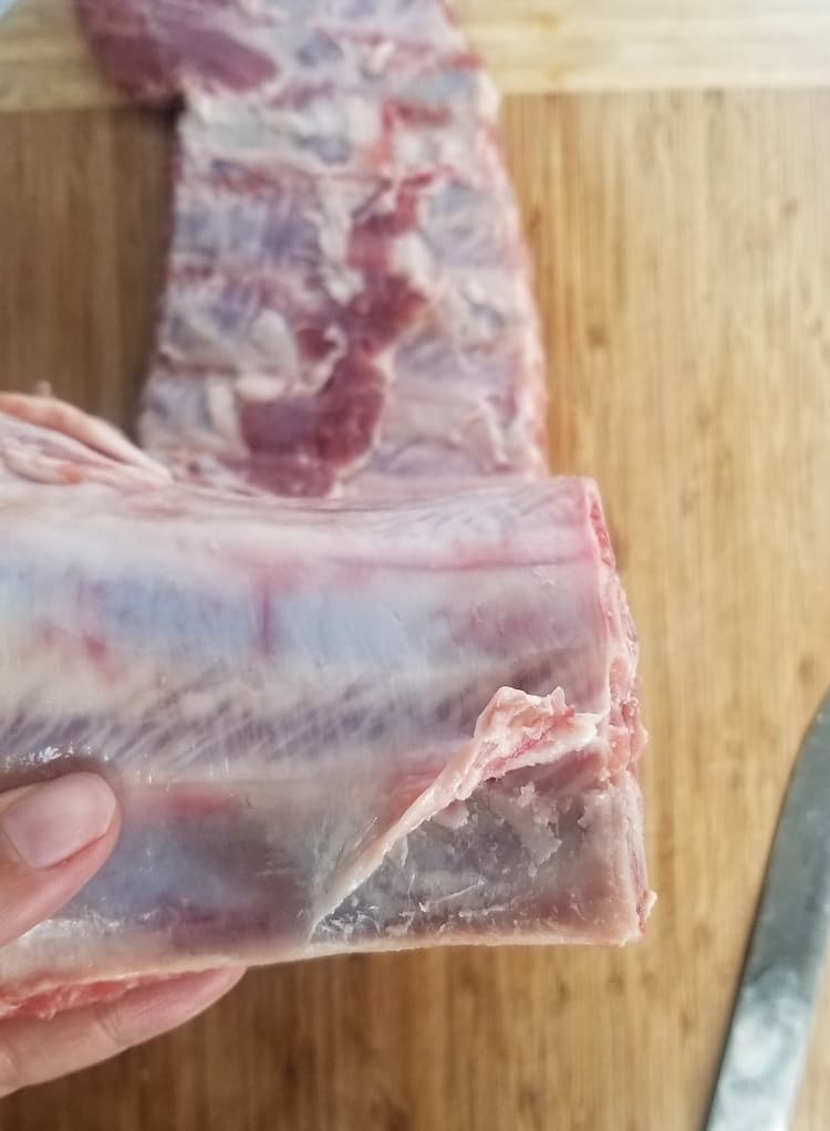 hand holding up the top of a rack of ribs to the camera showcasing a piece of the membrane lifted up
