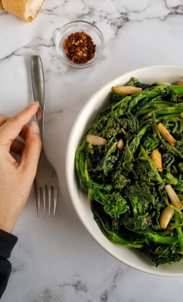 hand holding a fork next to a big bowl of sauteed rapini with garlic, bowl of chili flakes and slice of bread in the background