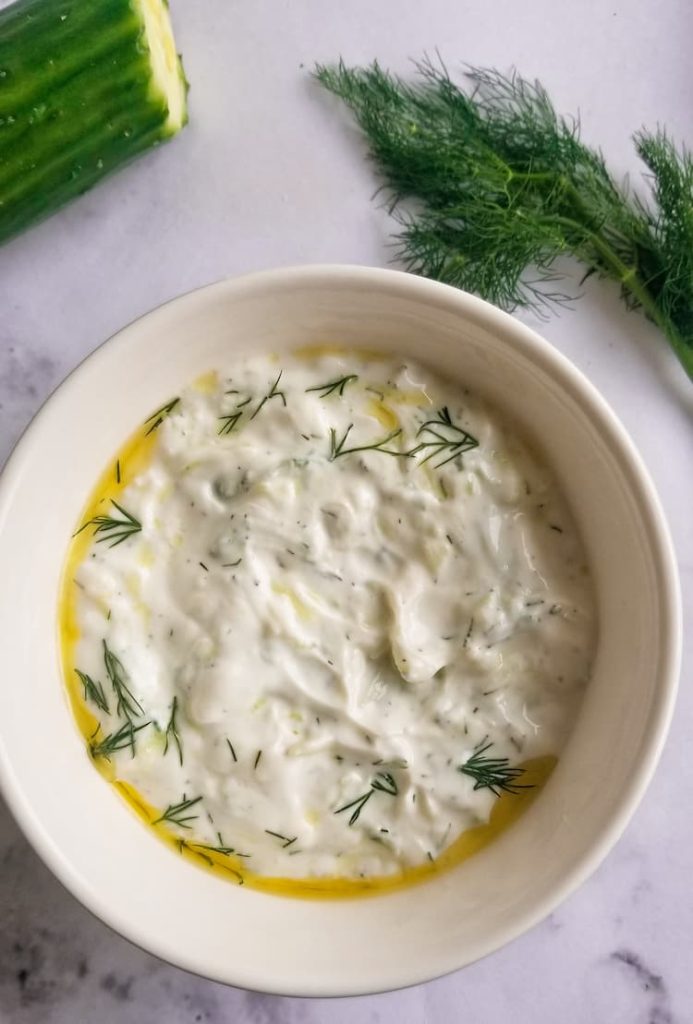 bowl of homemade tzatziki sauce with fresh dill on top and in the background, cut cucumber in the back, olive oil on top