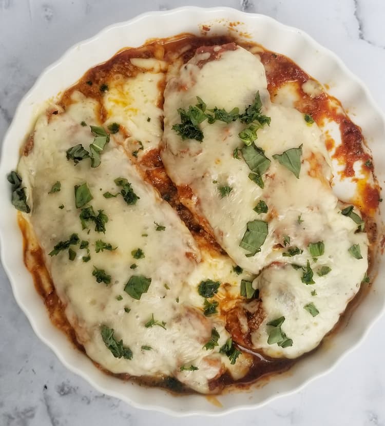 2 chicken breasts topped with melted mozzarella cheese, tomato sauce and fresh parsley in a white dish