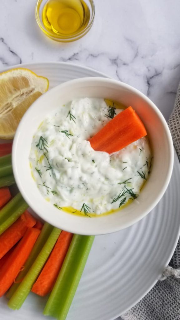 carrots, celery sticks and half a lemon on a white plate with a bowl of homemade tzatziki sauce, carrot dipped into the bowl, fresh dill and olive oil on top of the tzatziki, small bowl of olive oil behind
