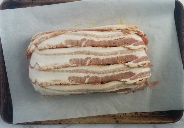 sheet pan with parchment paper with an uncooked meatloaf wrapped in raw bacon on top