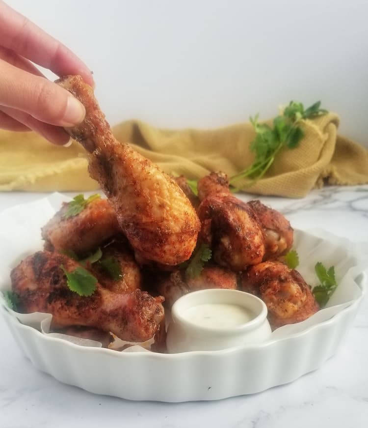 baked chicken drumsticks in a circular dish garnished with fresh parsley, creamy white sauce on the side with hand holding a drumstick over the sauce