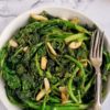 big bowl of sauteed rapini and garlic, two forks in the bowl, slices of bread in the background with a stemmed garlic bulb