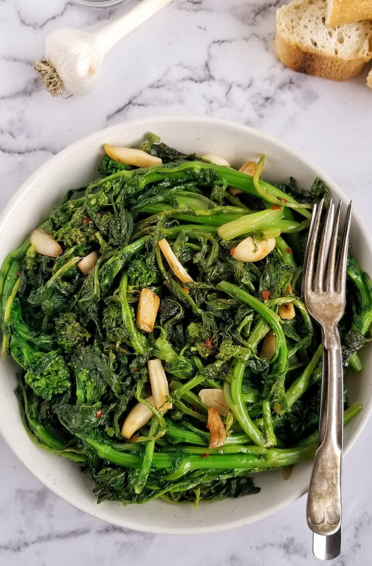 big bowl of sauteed rapini and garlic, two forks in the bowl, slices of bread in the background with a stemmed garlic bulb