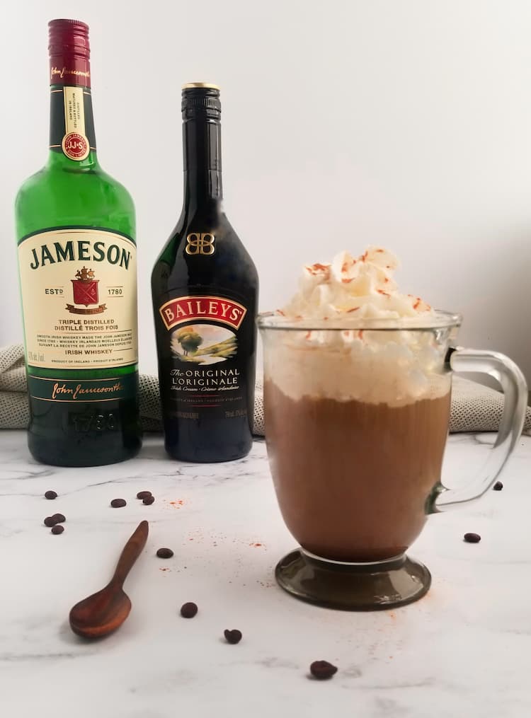 bottles of jameson and baileys next to a clear mug of irish coffee with baileys topped with whipped cream and cinnamon, wooden spoon and coffee beans in the front