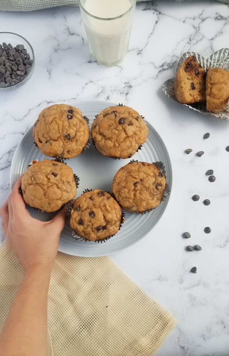glass of milk, bowl of chocolate chips, hand grabbing a freshly baked chocolate chip muffin off a plate with 4 other ones, split muffin in background, scattered chocolate chips on the right side