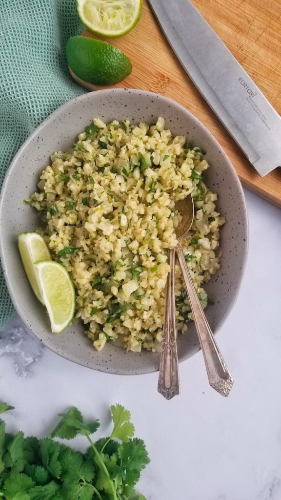 bowl of riced cauliflower mixed with cilantro, two limes wedges, two spoons in the bowl, cutting board i the back with more lime wedges and a knife. bunch of cilantro at the bottom