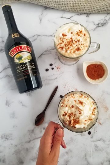 bottle of baileys laying next to two mugs, topped with whipped cream, hand holding the handle of one mug, bowl of cinnamon in the middle, coffee beans around, wooden spoon at the side