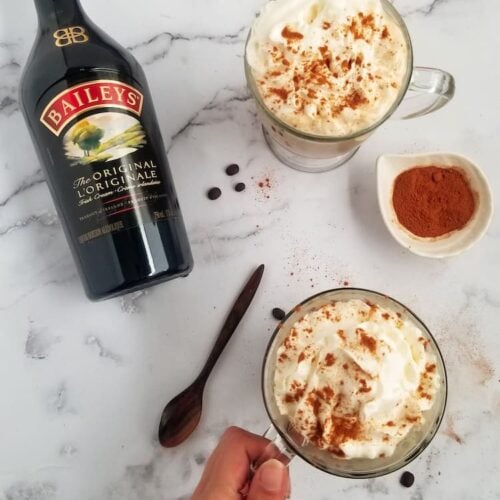 bottle of baileys laying next to two mugs, topped with whipped cream, hand holding the handle of one mug, bowl of cinnamon in the middle, coffee beans around, wooden spoon at the side.