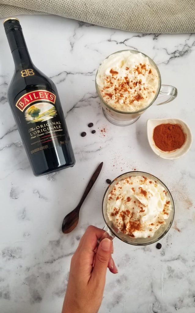 bottle of baileys laying next to two mugs, topped with whipped cream, hand holding the handle of one mug, bowl of cinnamon in the middle, coffee beans around, wooden spoon at the side