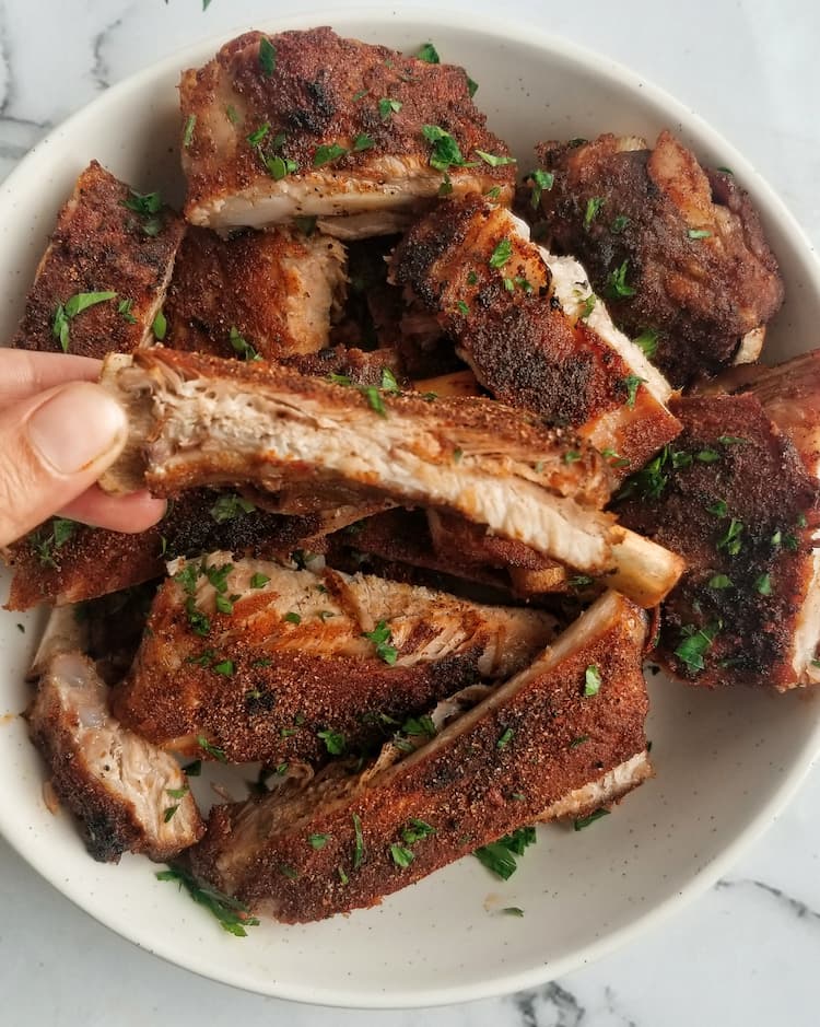 hand holding up a rib in front of bowl of cooked pork ribs topped with fresh chopped parsley