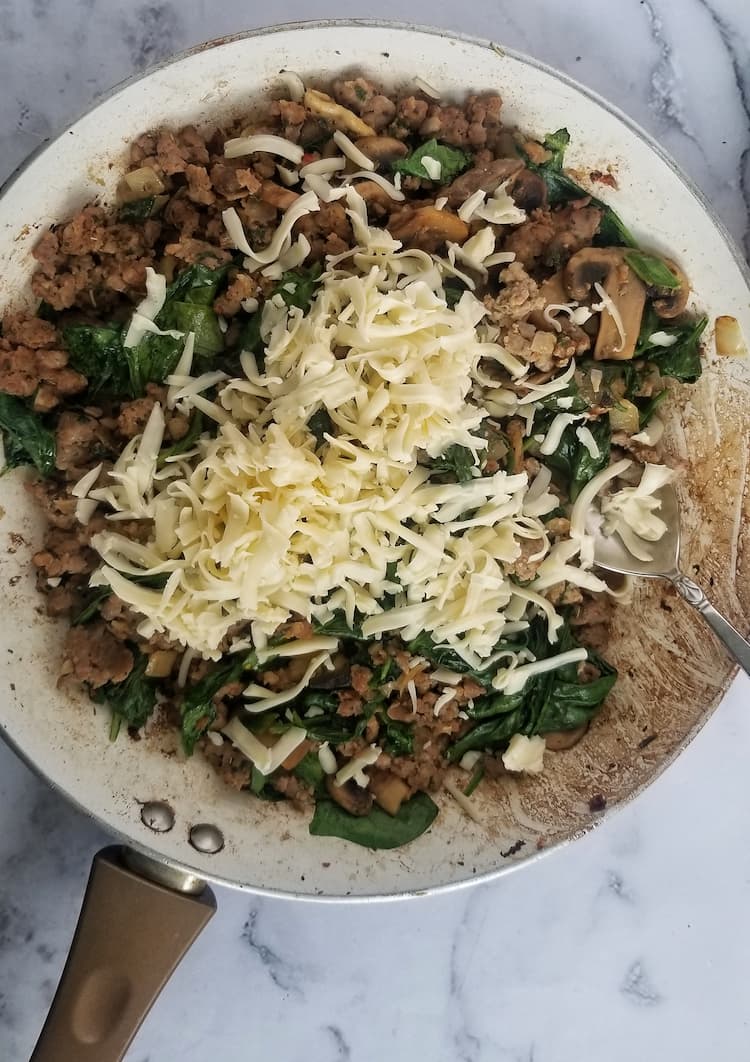 skillet with ground meat, shredded mozzarella, mushrooms, spinach and a silver spoon