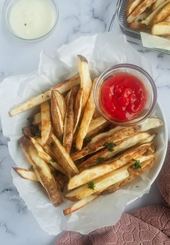 bowl of air fried french fries, topped with parsley, ramekin of ketchup in bowl at the top, tray of the rest of the fries behind beside a white dip