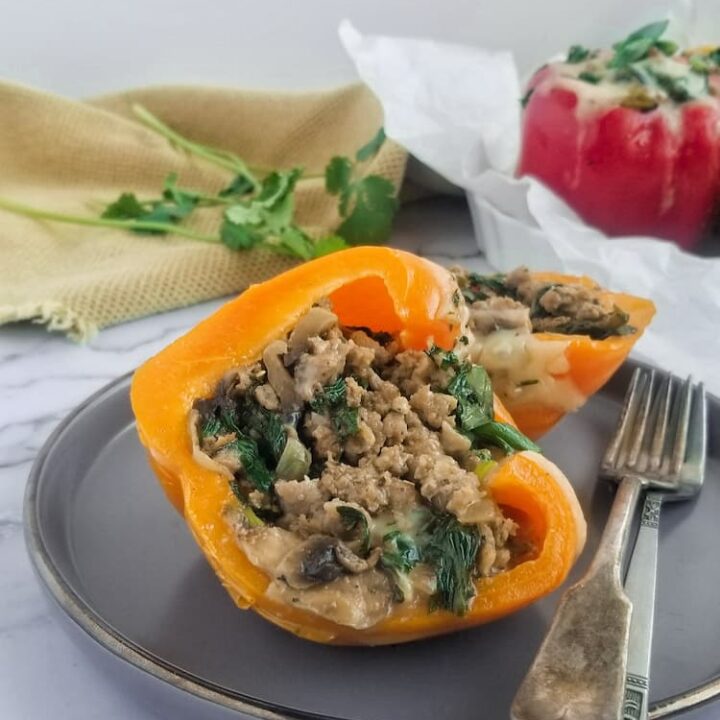 an orange stuffed pepper cut in half showcasing the ground meat, spinach and mushroom filling. 2 forks on the plate, red stuffed pepper in the background with some fresh parsley