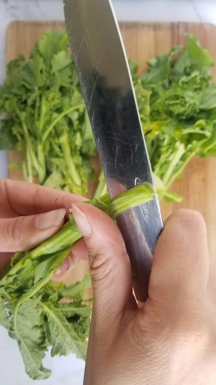 hand with a knife slicing the stem of a rapini over a bunch of the rest on a cutting board