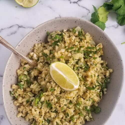 bowl of riced cauliflower mixed with cilantro, lime wedge in the middle on top and in the back, spoon in the bowl, bunch of cilantro in the back