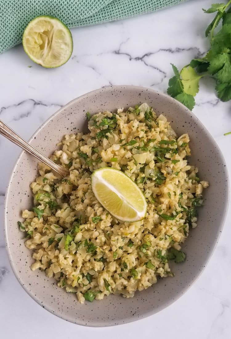 bowl of riced cauliflower mixed with cilantro, lime wedge in the middle on top and in the back, spoon in the bowl, bunch of cilantro in the back