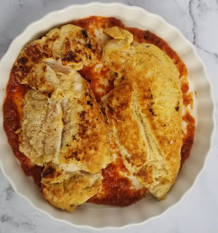 two breaded chicken breasts in a white dish sit on top of some tomato sauce