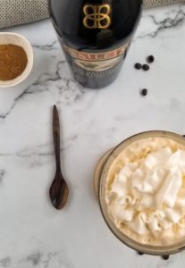 top view of glass mug of whipped cream, wooden spoon, bottle of baileys, ramekin of cinnamon and coffee beans