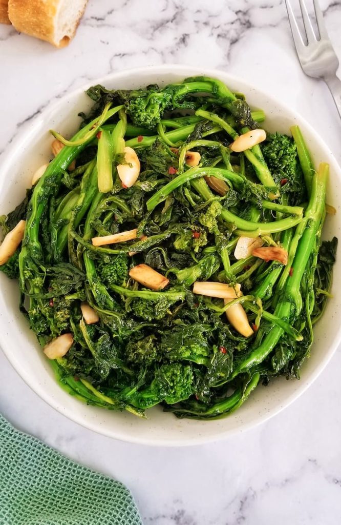 big bowl of sauteed rapini with garlic and chili flakes, fork on the top side and sliced bread of the top left